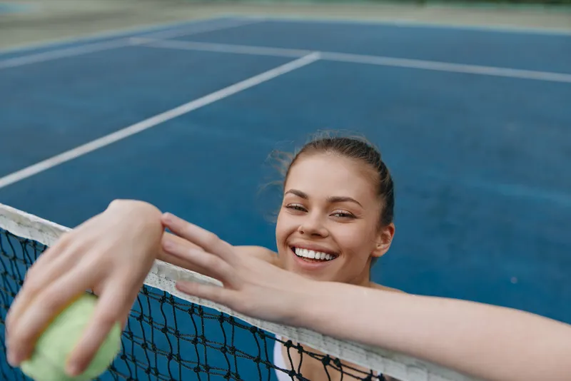 Young athletic female tennis player smiling while leaning on the net, showcasing a joyful expression and holding a tennis ball