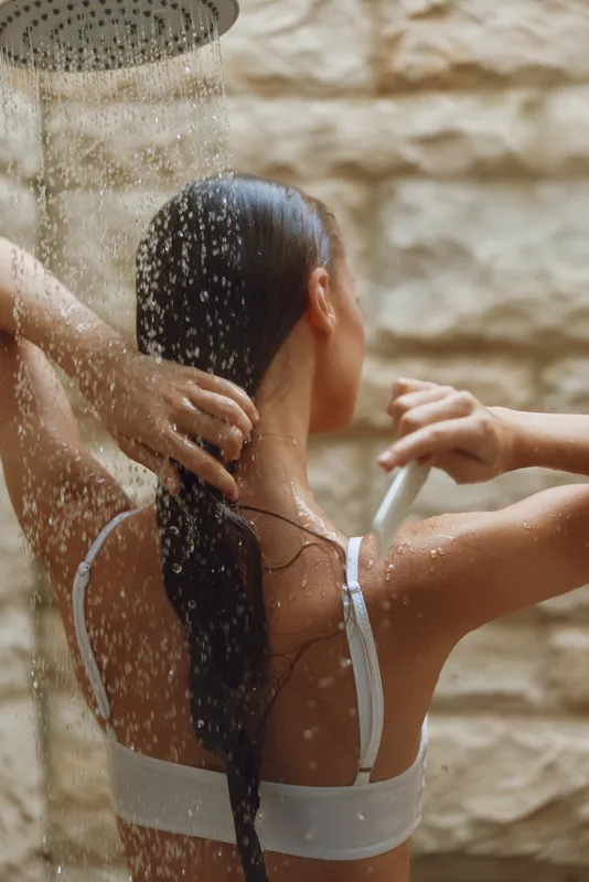 woman washing hair under shower with water droplets, wet skin, natural light, indoor bathroom setting, relaxing self-care moment