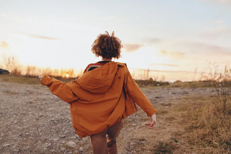 Woman lifestyle outdoors, seen from back, walking along a countryside path at sunset, wearing a warm jacket, arms relaxed, golden light, calm mood, soft wind, natural colors