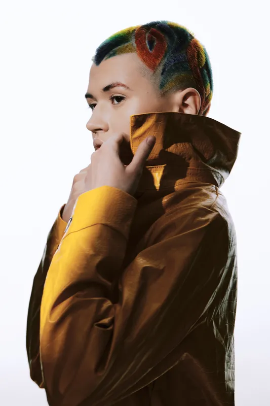 young woman with colorful patterned hair wearing yellow jacket posing thoughtfully against white background, creative hairstyle and fashion style