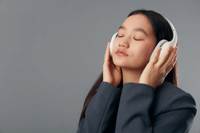 Young woman with long hair wearing headphones, eyes closed, enjoying music and relaxing with calm expression in studio with gray background and casual dark jacket.