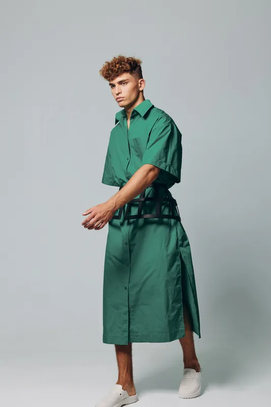 Young man with curly hair wearing green oversized shirt dress and white slip-on shoes posing in studio with neutral background, fashion and style concept.