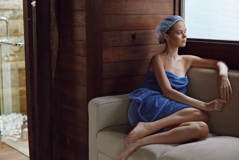 Relaxed woman in spa robe and hair cap sitting on beige sofa in wooden interior, enjoying calm moment with soft natural light and peaceful atmosphere indoors.