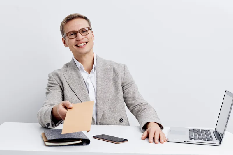 Professional businessman in smart casual attire smiling at camera while holding a document and working on his laptop sitting at a desk in modern office environment. Business, success, confident, workp
