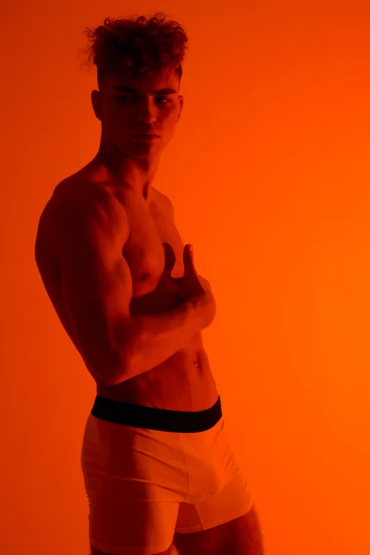 Muscular man posing shirtless in white shorts with dramatic orange lighting, showing defined abs and strong physique in studio for fitness and health concept.