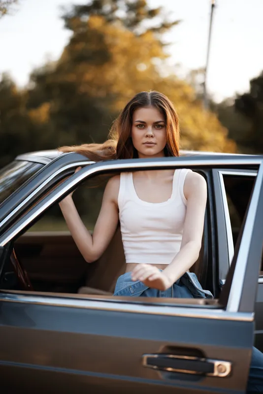 Young woman in casual summer attire leaning out of a car, with a serene expression, surrounded by greenery, embodying freedom and adventure.