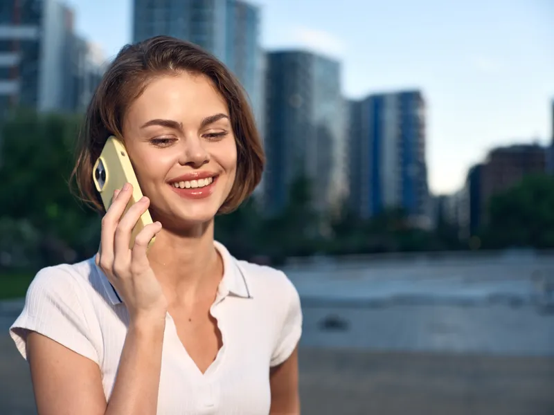 A cheerful business professional smiles while talking on a mobile phone outdoors in a modern urban setting, conveying optimistic, confident energy during a sunny day by the city.