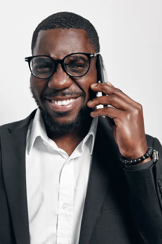 African man with dark skin wearing glasses and black suit talking on smartphone smiling in studio. Business professional, communication, technology and confident male portrait with white background.