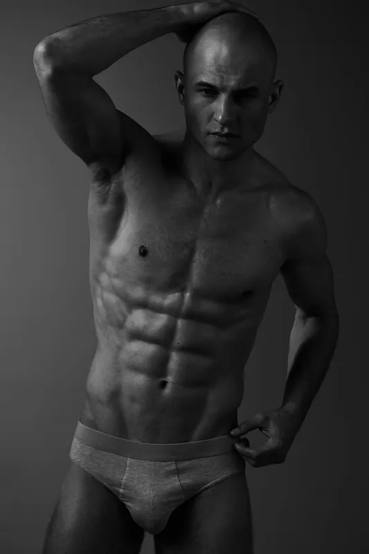 Black and white portrait of a muscular man striking a pose, showcasing his toned physique and confidence in minimal clothing.