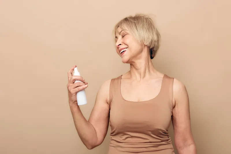 Smiling mature woman with short blonde hair sprays facial mist on her face in a beige tank top. Studio shot with soft lighting on neutral background. Concept of skincare, hydration, and self-care.