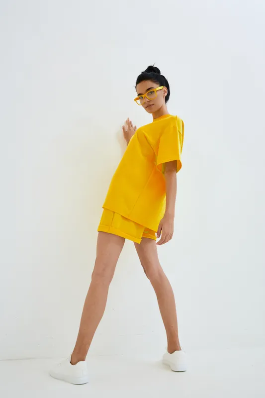 Young woman in bright yellow casual outfit posing against white background, wearing stylish glasses and white sneakers, showcasing modern minimalistic fashion.
