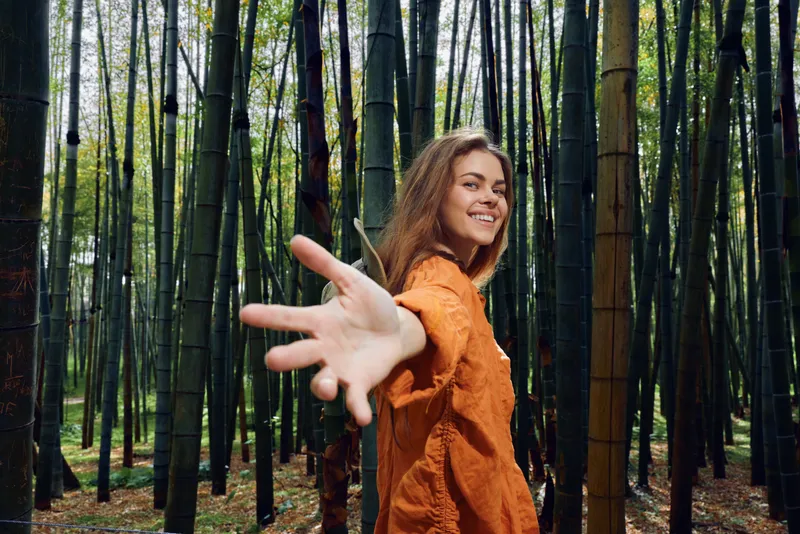 woman bamboo forest smile hand reach nature portrait: smiling young woman in orange shirt reaches her hand toward the camera in a dense bamboo grove, outdoor travel and joyful connection.
