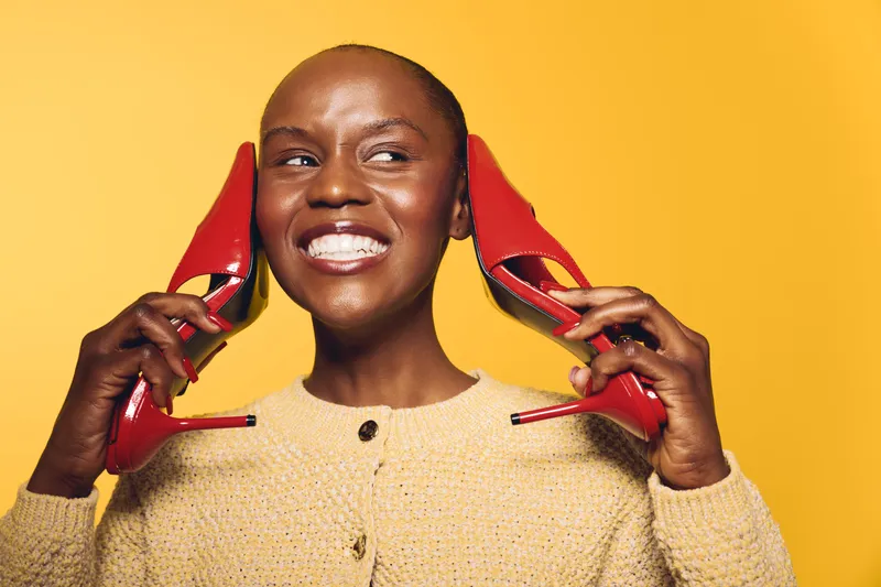 Smiling dark skinned African woman holds red high heels near ears like phones, wearing beige sweater. Bright yellow background highlights joyful expression and playful mood in studio portrait.