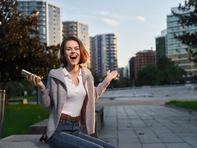 A cheerful young woman sits outdoors in an urban park area with tall buildings in the background. She smiles brightly, arms open, enjoying a sunny day.