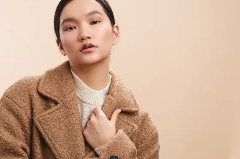 Asian woman in a warm studio setting wears a tan coat and light sweater. Her calm, confident gaze and soft lighting create a stylish, contemporary portrait with inviting texture.