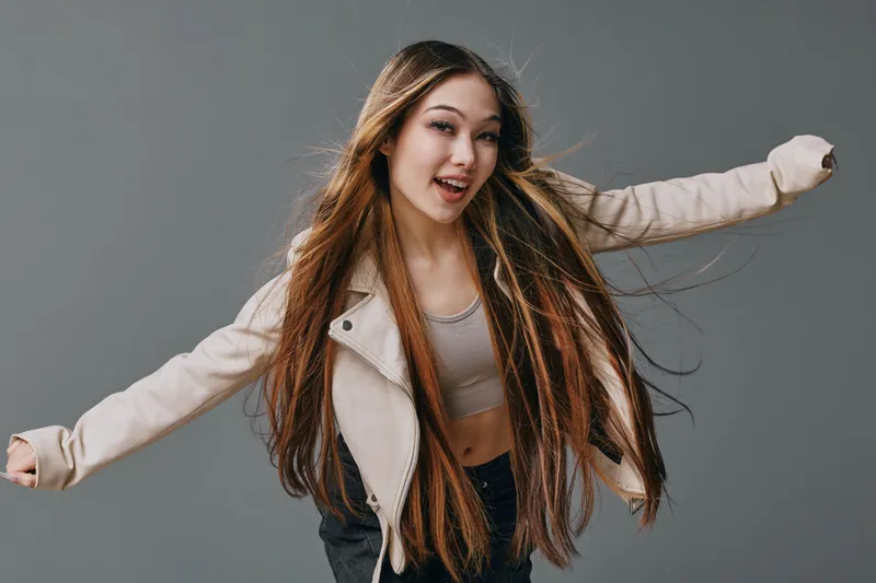 Young woman with long hair wearing casual jacket and crop top posing and smiling in studio, expression of joy and freedom with natural light and gray background