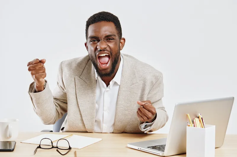 Confident happy young African-American man in beige blazer showing fist gesture and smiling with excitement, sitting at desk with laptop, glasses, and stationery on white background, studio portrait. 