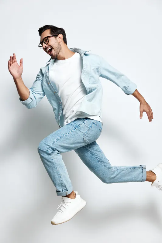 Energetic smiling man in casual outfit jumps joyfully against plain light background, showcasing excitement and happiness. Lifestyle and freedom concept.
