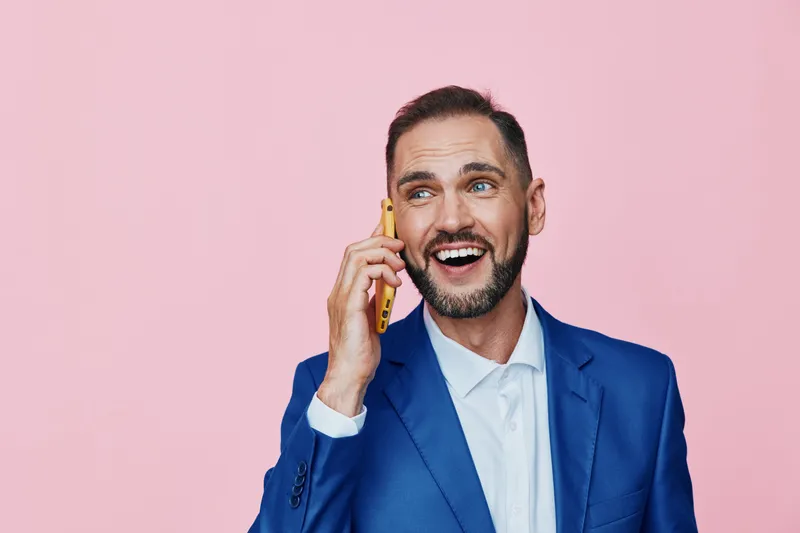 Professional man in a blue suit on a solid pink background, engaged in a phone call, showing confidence, enthusiasm, and friendly energy in a corporate setting, for business media use across channels.