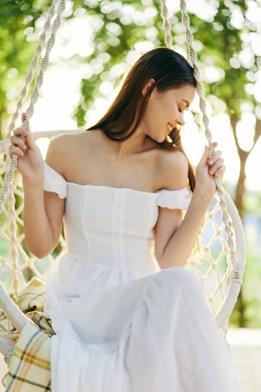Elegant young woman in a white dress enjoying a sunny day on a swing in a lush green park, radiating joy and tranquility