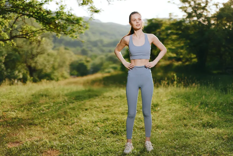 Young woman in sportswear smiling outdoors in a sunny green landscape, promoting fitness and healthy lifestyle concepts