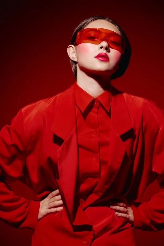 Confident woman wearing oversized red blazer and vivid red sunglasses poses with hands on hips. Fashion-forward style expresses creativity and bold trends in beauty styling.