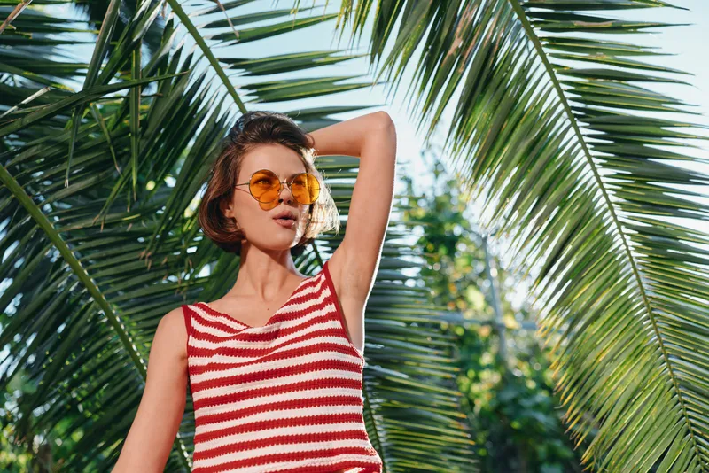 A stylish woman poses outdoors under lush palm leaves, wearing a red striped tank top and round sunglasses. She exudes confidence, sunshine, and casual chic.