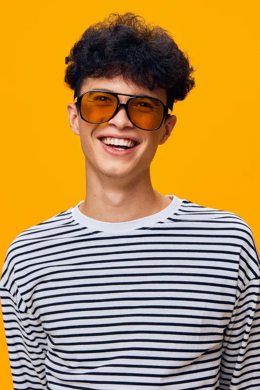 young man smiling wearing yellow sunglasses and striped shirt, curly hair, bright orange backdrop, cheerful expression, casual style