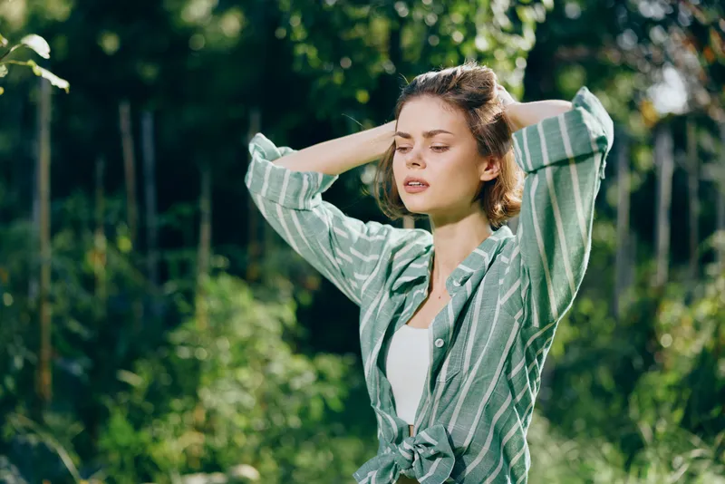 A woman stands outdoors in a sunlit park, wearing a striped green shirt with sleeves rolled, hands behind her head, and a relaxed, calm expression.