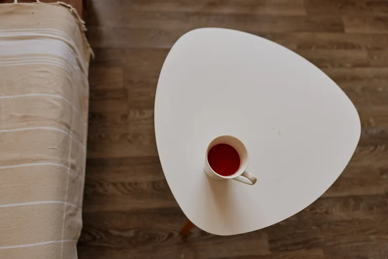 A cozy home lifestyle scene with a round white table beside a bed, a small cup of tea on the surface, and warm natural light. Clear concept of comfort, calm, and everyday home life.