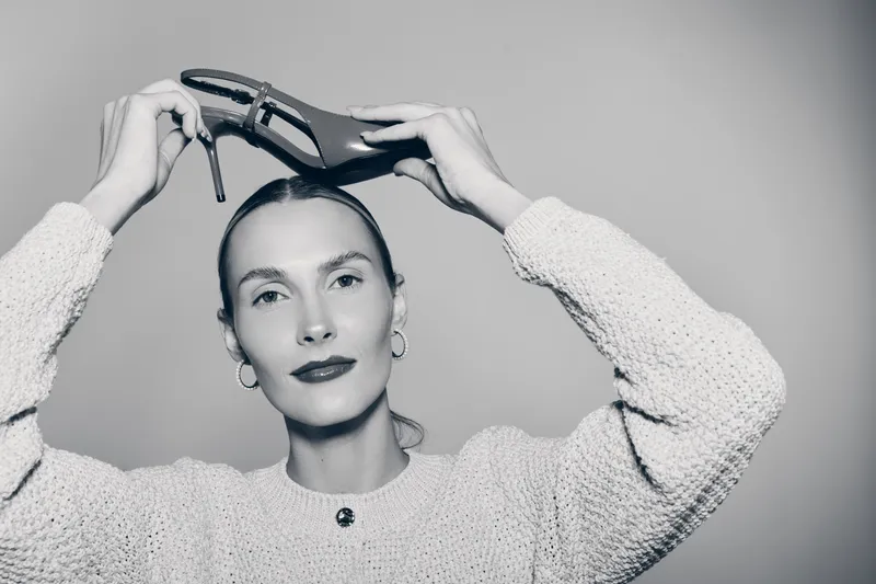 Slavic woman with fair skin holds high heel shoe above head, posing in a knitted sweater against grey background. Concept for fashion, style, and minimalism.