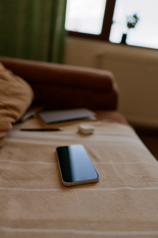 A quiet home scene with a smartphone resting on a bed, soft natural light from a window, papers nearby. A woman lifestyle moment capturing calm routine and everyday technology.