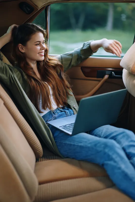 Happy young woman with long hair using laptop in a car, wearing casual outfit, enjoying a relaxing moment in nature, vibrant colors, modern lifestyle.