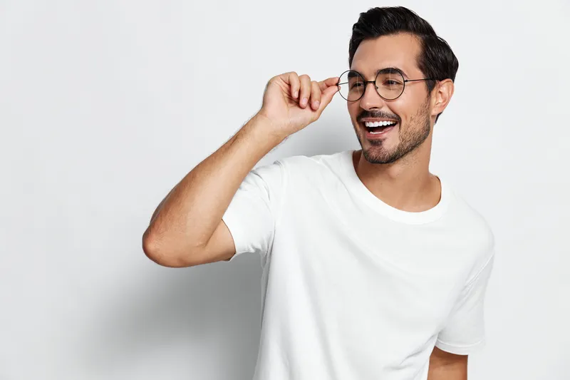 Smiling young man in white t-shirt adjusting his glasses and looking at the camera. Fun, cheerful mood captured in a studio with a clean background.