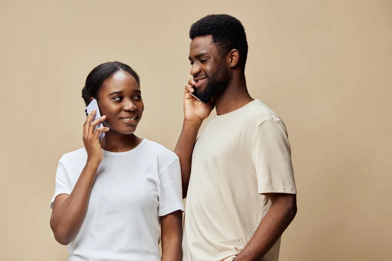 A happy Black couple enjoying a phone conversation, demonstrating connection and joy, dressed casually in neutral colors on a beige background