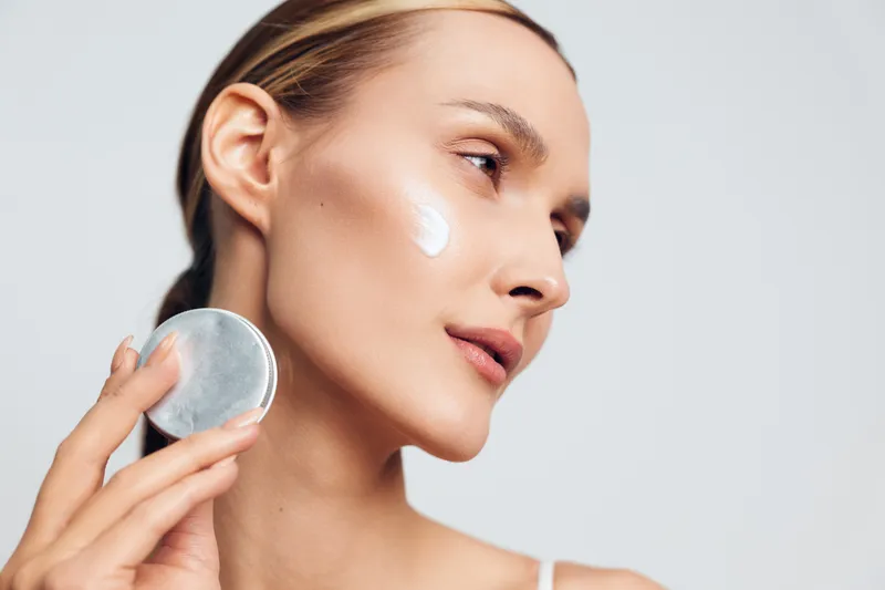 Young woman applies moisturizing cream on her face with a cosmetic sponge, skincare routine concept, healthy skin, natural beauty, isolated on white background.