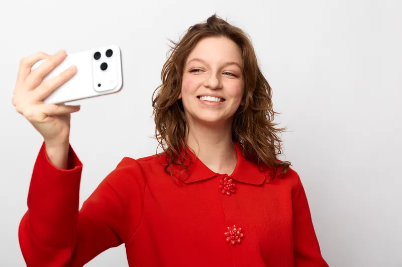 Smiling young woman with curly hair wearing red sweater takes selfie with smartphone on white background. Concept of social media, communication, and casual lifestyle.