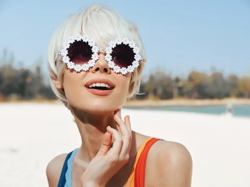 Portrait of a stylish woman with platinum hair wearing oversized flower sunglasses on a sunny beach, capturing bold fashion and a carefree summer mood.