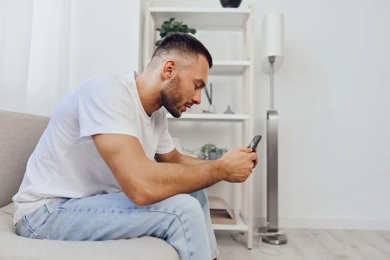 Pensive man sitting on the couch, experiencing digital detox while engaging in emotional self-reflection and managing work-life blur with technology in hand.