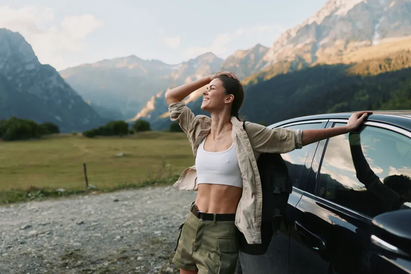 Young woman in casual clothes leans on a car and enjoys a mountain landscape with closed eyes and relaxed expression during a sunny day outdoors.