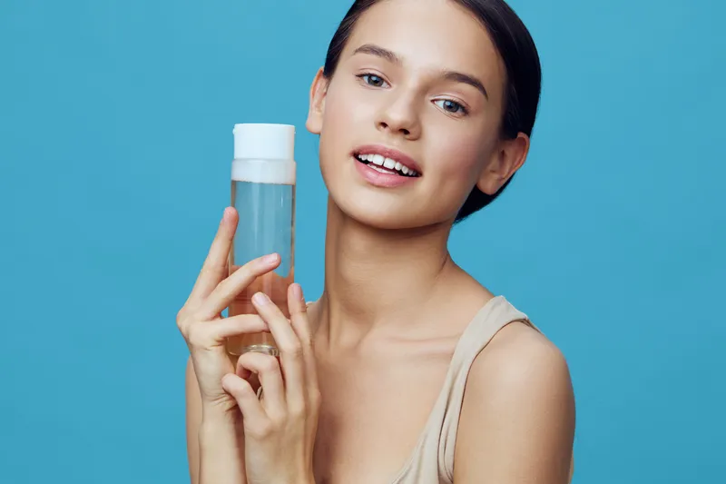 Young woman holding a clear skincare toner bottle, smiling with a healthy glow against a light blue background, representing beauty and self care