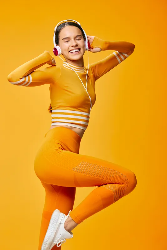Fitness woman in bright orange athletic wear jumping with headphones on, midair pose against a vivid orange background, showcasing energy, movement and sport lifestyle.