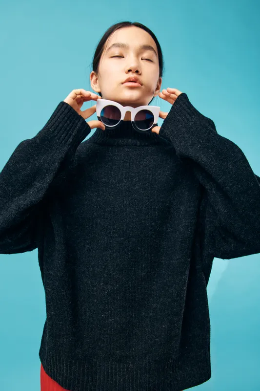 Young woman wearing black sweater holding white sunglasses near neck standing against blue background with closed eyes fashion style casual outfit comfortable clothes autumn season.