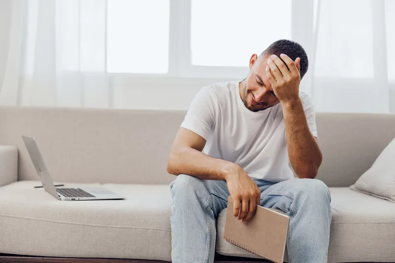 Thoughtful man sitting on a couch, reflecting while holding a notebook, with a laptop nearby, showcasing a modern and cozy living space atmosphere.