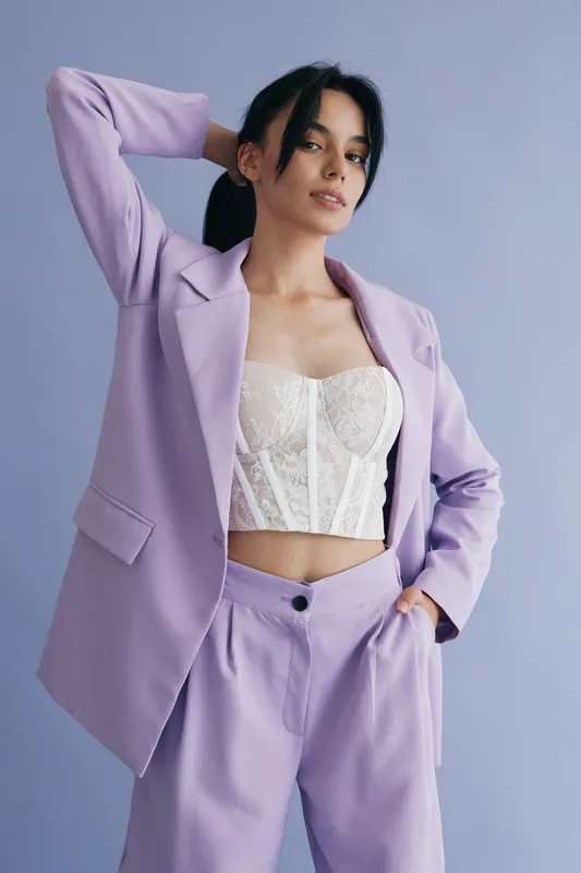 Young woman in lilac suit and white lace corset posing confidently against soft lavender background, showcasing modern fashion style and feminine elegance.