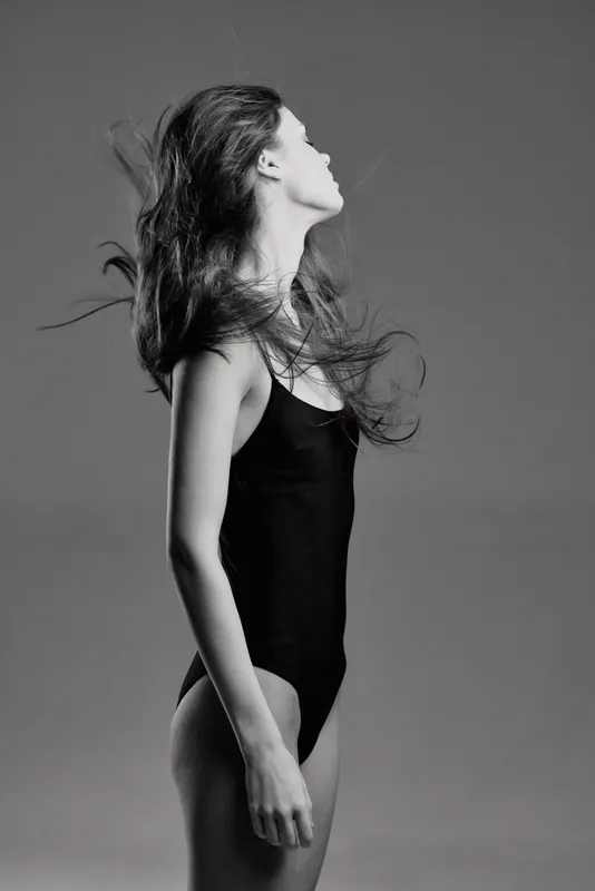 Black and white portrait of a woman with flowing hair wearing a black bodysuit in studio lighting.