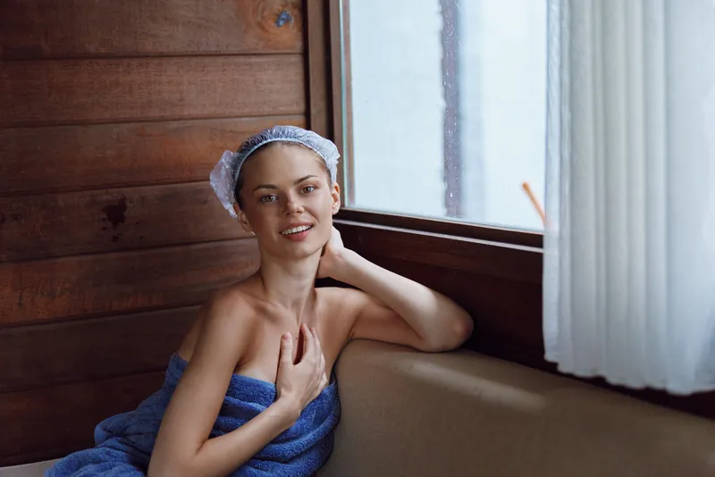 Smiling woman wrapped in blue towel relaxing indoors near wooden window with soft natural light and sheer white curtains, enjoying peaceful moment after bath.