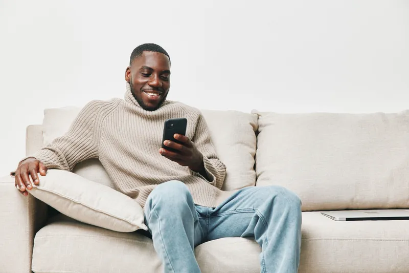 Smiling young man with a smartphone relaxing on a cozy couch in casual attire, conveying a happy and relaxed mood Key elements include comfort and modern lifestyle