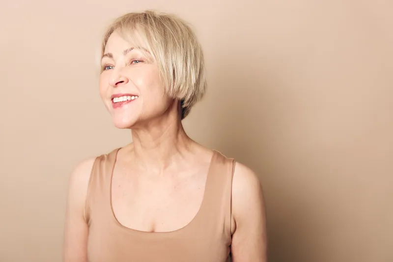 Smiling middle aged woman with short blonde hair stands against beige background wearing sleeveless top. Natural beauty concept with soft lighting highlighting joyful expression and healthy skin.