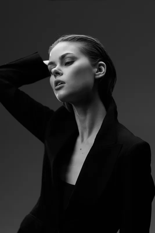 Black and white portrait of a stylish woman with closed eyes wearing a dark blazer, expressing calm and elegance in minimalistic lighting.